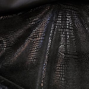 May include: Close-up of a black fabric with a reptile-like texture. The material has a sheen, with light reflecting off the surface, creating highlights and shadows. The fabric is draped, showing folds and curves.
