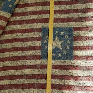 May include: A woven fabric featuring a repeating pattern of red and cream horizontal stripes with blue squares containing a large central star surrounded by smaller stars. A yellow measuring tape shows a length of approximately 89 centimetres.
