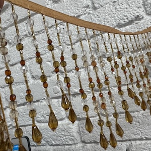 May include: A decorative beaded fringe with a tan backing. The fringe features clear, amber, and brown beads in various shapes, including round and teardrop. The beads are strung together with gold-colored thread, creating a shimmering effect.