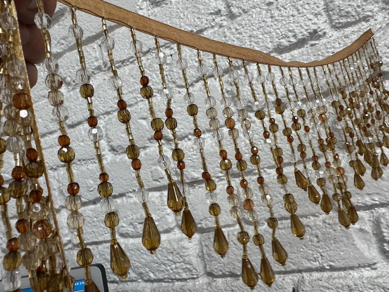 May include: A decorative beaded fringe with a tan backing. The fringe features clear, amber, and brown beads in various shapes, including round and teardrop. The beads are strung together with gold-colored thread, creating a shimmering effect.