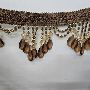 May include: Brown decorative trim with beaded accents and teardrop-shaped crystal pendants. The trim features a woven design with intricate details and is suitable for various craft projects.