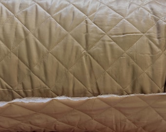 May include: Close-up of a rolled-up, quilted fabric in a warm, golden-tan color. The fabric features a diamond pattern created by stitched lines. The edge of the fabric reveals a fluffy, white material, possibly batting or padding, adding texture and dimension.