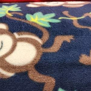 May include: A navy blue fleece blanket featuring a repeating pattern of smiling cartoon monkeys swinging from brown branches with green and yellow leaves. The monkeys have white faces and brown bodies.
