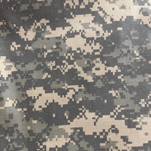 Op de afbeelding: Een close-up van een camouflage stof met een gepixeld patroon in tinten bruin, groen en grijs.