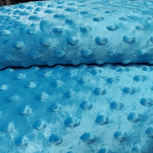 K&ouml;nnte beinhalten: Nahaufnahme eines weichen, blauen Stoffes mit einem strukturierten, gepunkteten Muster. Der Stoff ist in der Mitte gefaltet, wodurch die Textur auf beiden Seiten sichtbar wird.
