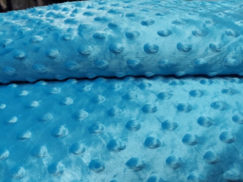 K&ouml;nnte beinhalten: Nahaufnahme eines weichen, blauen Stoffes mit einem strukturierten, gepunkteten Muster. Der Stoff ist in der Mitte gefaltet, wodurch die Textur auf beiden Seiten sichtbar wird.