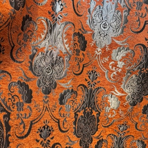 May include: Close-up of a textured fabric with an intricate damask pattern. The design features a vibrant orange background with detailed gray and black floral and scroll motifs. The fabric has a luxurious appearance.