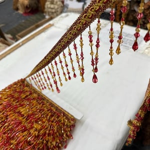 May include: A decorative trim featuring a woven gold and red fabric header with dangling strands of faceted red and amber teardrop beads. The trim is arranged in a dense, layered pile of beaded fringe, showcasing a repeating pattern of beads.