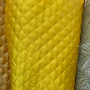 May include: A roll of bright yellow quilted fabric with a diamond pattern. The fabric has a shiny, satin-like appearance. The quilting creates a textured surface. Other fabrics are visible in the background.
