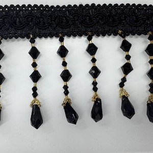 May include: Black beaded fringe trim with a black woven top. The fringe is made of faceted black beads with gold accents.