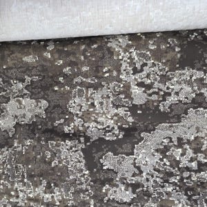 May include: A rolled-up section of patterned fabric. The fabric features a dark gray and brown base with a speckled, abstract design in shades of silver and white. The top edge shows a rolled section of plain white fabric.
