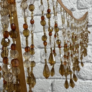 May include: A decorative beaded curtain with a wooden top and strands of amber, clear, and gold beads. The beads are various shapes and sizes, including round and teardrop. The curtain hangs in front of a white brick wall.