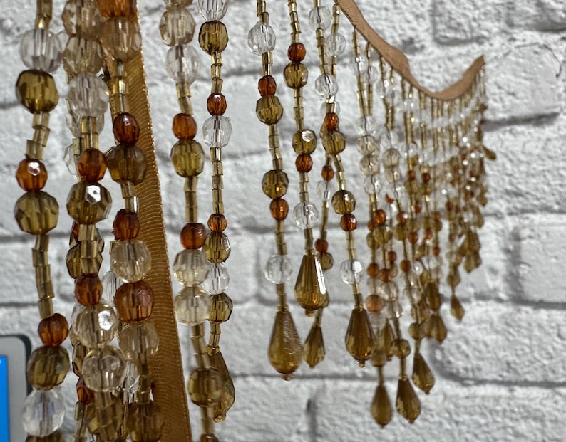May include: A decorative beaded curtain with a wooden top and strands of amber, clear, and gold beads. The beads are various shapes and sizes, including round and teardrop. The curtain hangs in front of a white brick wall.