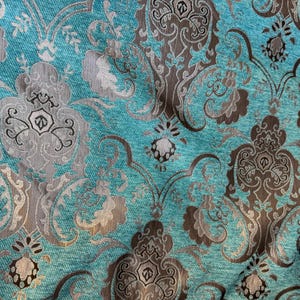 Op de afbeelding: Close-up van een turquoise stof met een damastpatroon. Het ontwerp bevat sierlijke, zich herhalende bloemen- en scrollmotieven in grijstinten en bruin. De stof heeft een getextureerd uiterlijk, wat wijst op een geweven materiaal.