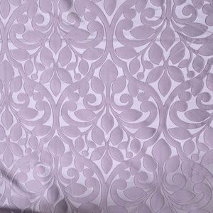 May include: A close-up of a lavender fabric with a repeating floral pattern. The design features stylized leaves and swirling lines, creating a textured, elegant look. The fabric has a subtle sheen, suggesting a luxurious material.