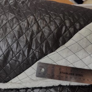 May include: Close-up of black quilted fabric with a diamond pattern. The fabric is shiny and has a ruler with inch measurements laid across it. The ruler reads up to 76 cm.