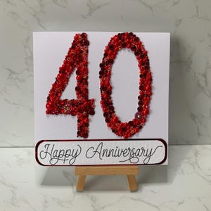 May include: A white card with the number "40" in red beads and the words "Happy Anniversary" in script. The card is displayed on a small wooden easel, perfect for a milestone celebration.