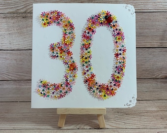 May include: A white card with the number 30 made of colorful flowers. The number 30 is decorated with a small red ladybug.