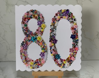 Puede incluir: Una tarjeta blanca con borde festoneado presenta el número 80 hecho de cuentas y pedrería de colores. La tarjeta se muestra en un pequeño caballete de madera.