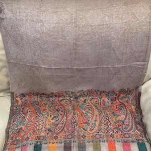 May include: A rectangular scarf with a light brown background and a detailed, colorful border. The border features a paisley design in shades of orange, blue, green, and purple. The scarf has a striped fringe at the bottom.