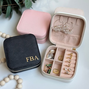 May include: Three square jewelry cases in black, pink, and white. The black case features the gold letters FBA. The open white case reveals a light pink interior with compartments for rings, earrings, and necklaces, containing various jewelry pieces.