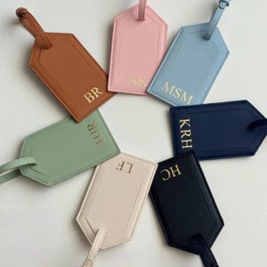 Puede incluir: Un conjunto de etiquetas de equipaje personalizadas en varios colores: marr&oacute;n, rosa, azul, verde, beige, azul marino y negro. Cada etiqueta tiene iniciales doradas y una correa a juego. Las etiquetas est&aacute;n dispuestas en c&iacute;rculo.