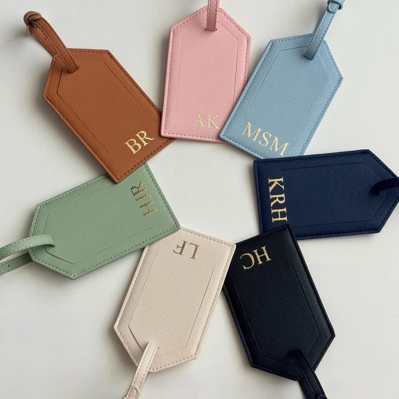 Puede incluir: Un conjunto de etiquetas de equipaje personalizadas en varios colores: marr&oacute;n, rosa, azul, verde, beige, azul marino y negro. Cada etiqueta tiene iniciales doradas y una correa a juego. Las etiquetas est&aacute;n dispuestas en c&iacute;rculo.