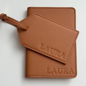 May include: A tan passport holder and a matching luggage tag, both featuring a textured finish and the name LAURA embossed in capital letters. The luggage tag is attached to the passport holder with a thin strap, and both items are tan.