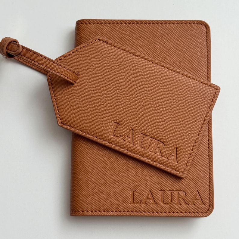 May include: A tan passport holder and a matching luggage tag, both featuring a textured finish and the name LAURA embossed in capital letters. The luggage tag is attached to the passport holder with a thin strap, and both items are tan.