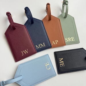 Puede incluir: Una colecci&oacute;n de etiquetas de equipaje personalizadas en varios colores, incluyendo burdeos, azul marino, marr&oacute;n, verde salvia, azul claro y negro. Cada etiqueta presenta iniciales doradas y una correa a juego.
