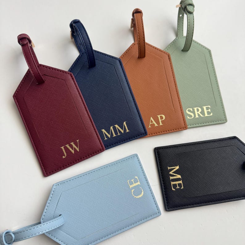 Puede incluir: Una colecci&oacute;n de etiquetas de equipaje personalizadas en varios colores, incluyendo burdeos, azul marino, marr&oacute;n, verde salvia, azul claro y negro. Cada etiqueta presenta iniciales doradas y una correa a juego.