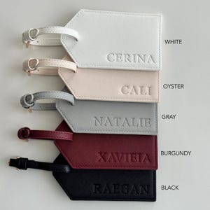 May include: Five rectangular luggage tags with buckled straps, each featuring a textured finish and a debossed name. The colors from top to bottom are white with CERINA, oyster with CALI, gray with NATALIE, burgundy with XAVIEIA, and black with RAEGAN.