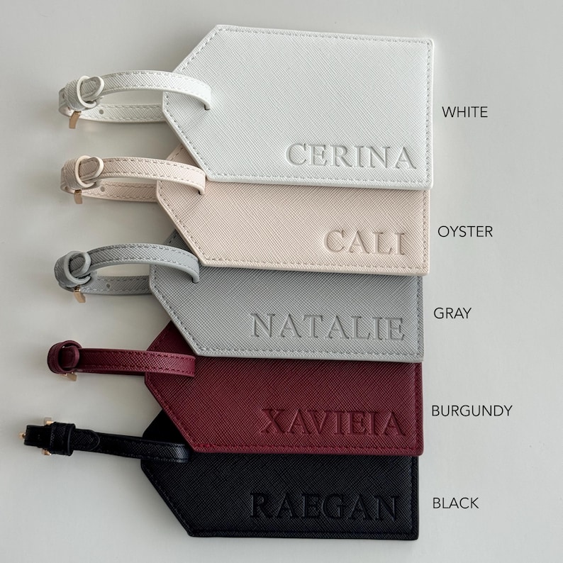 May include: Five rectangular luggage tags with buckled straps, each featuring a textured finish and a debossed name. The colors from top to bottom are white with CERINA, oyster with CALI, gray with NATALIE, burgundy with XAVIEIA, and black with RAEGAN.