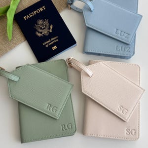 May include: A dark blue United States passport next to three sets of passport holders and luggage tags in light blue, sage green, and pale pink. Each holder and tag is embossed with initials LUZ, RG, or SG in a clean, sans-serif font.