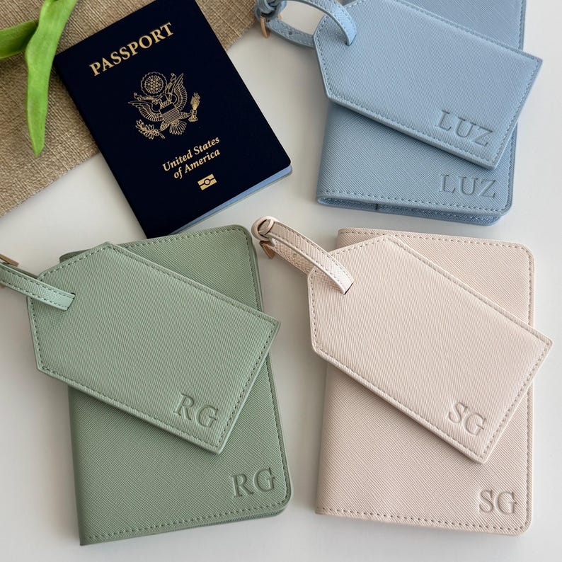 May include: A dark blue United States passport next to three sets of passport holders and luggage tags in light blue, sage green, and pale pink. Each holder and tag is embossed with initials LUZ, RG, or SG in a clean, sans-serif font.
