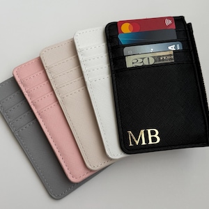 May include: Five card holders in different colors, including gray, pink, beige, white, and black. The black card holder has a zipper and the initials "MB" in gold lettering.