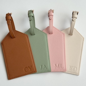 May include: Four personalized luggage tags in shades of brown, green, pink, and cream. Each tag has a strap and buckle at the top and features embossed initials. The tags are arranged in a row, showcasing a variety of colors and designs.