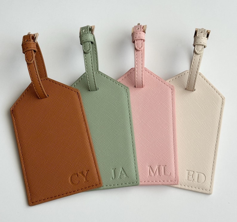 May include: Four personalized luggage tags in shades of brown, green, pink, and cream. Each tag has a strap and buckle at the top and features embossed initials. The tags are arranged in a row, showcasing a variety of colors and designs.