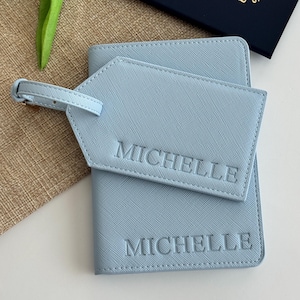 May include: A light blue passport holder and a matching luggage tag, both featuring a textured finish and the name MICHELLE embossed in capital letters. The luggage tag includes a small buckled strap for attachment to a suitcase or bag.