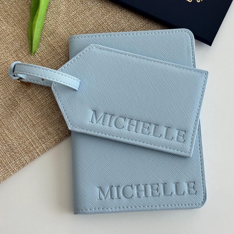 May include: A light blue passport holder and a matching luggage tag, both featuring a textured finish and the name MICHELLE embossed in capital letters. The luggage tag includes a small buckled strap for attachment to a suitcase or bag.