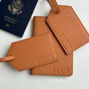 May include: A cognac-colored travel set featuring a passport holder, two luggage tags, and a passport. The luggage tags and passport holder are embossed with the name "LUCY". The passport is navy blue and reads "United States of America".