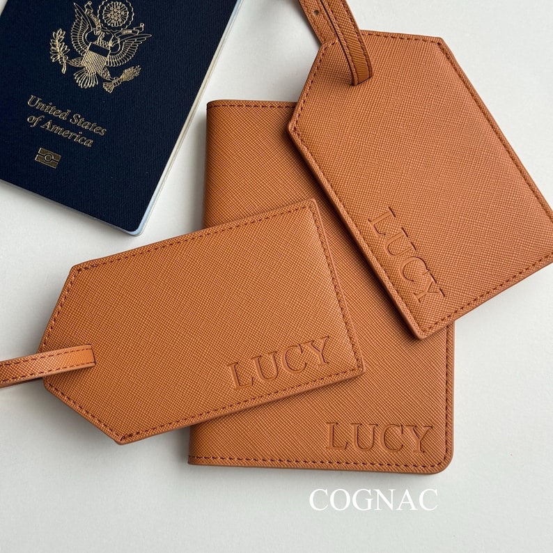 May include: A cognac-colored travel set featuring a passport holder, two luggage tags, and a passport. The luggage tags and passport holder are embossed with the name "LUCY". The passport is navy blue and reads "United States of America".