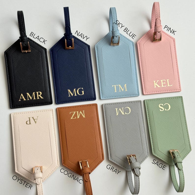 Puede incluir: Una colecci&oacute;n de etiquetas de equipaje personalizadas en varios colores, incluyendo negro, azul marino, azul cielo, rosa, ostra, co&ntilde;ac, gris y salvia. Cada etiqueta presenta iniciales doradas y el nombre del color. Las etiquetas tienen una hebilla y una correa.