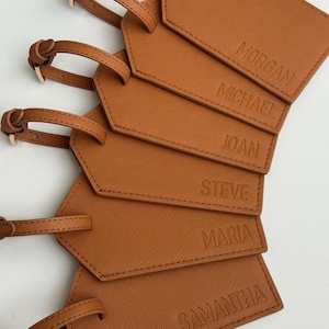May include: A stack of brown luggage tags with the names Morgan, Michael, Joan, Steve, Maria, and Samantha embossed on them. Each tag has a matching brown strap with a gold-colored buckle.