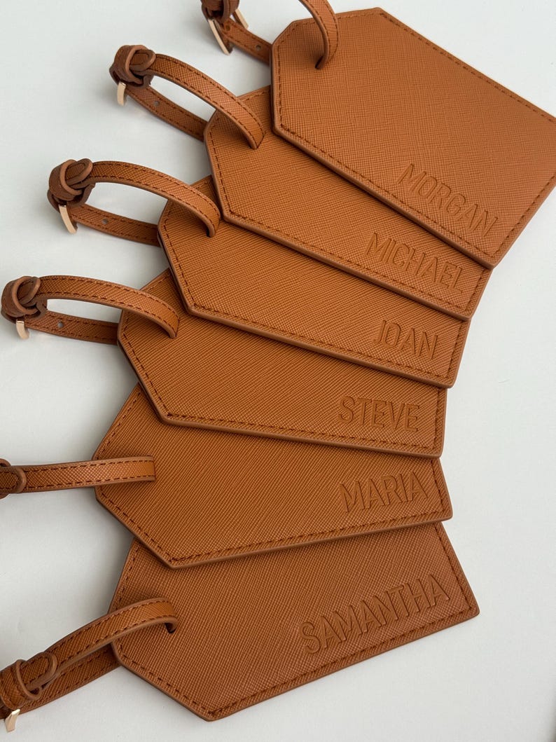 May include: A stack of brown luggage tags with the names Morgan, Michael, Joan, Steve, Maria, and Samantha embossed on them. Each tag has a matching brown strap with a gold-colored buckle.