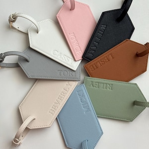 May include: A collection of personalized luggage tags in various colors, including white, pink, black, brown, gray, beige, blue, and green. Each tag features a name embossed in a matching tone, with a strap for attachment.