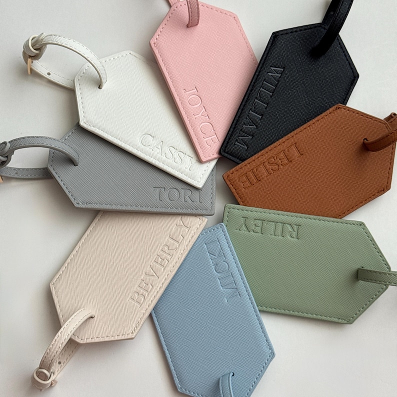 May include: A collection of personalized luggage tags in various colors, including white, pink, black, brown, gray, beige, blue, and green. Each tag features a name embossed in a matching tone, with a strap for attachment.