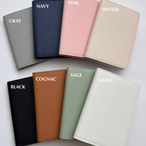May include: A collection of passport covers in various colors, including gray, navy, pink, oyster, black, cognac, sage, and white. Each cover is labeled with its color name. The text "PASSPORT COVERS" is at the bottom.