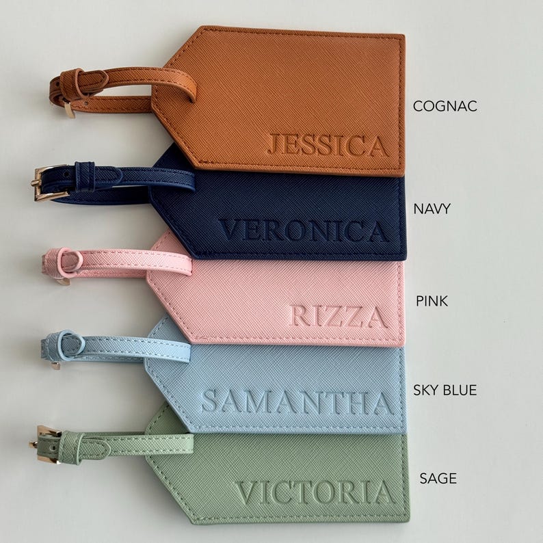 May include: Five rectangular luggage tags with buckled straps, each featuring a textured surface and embossed names. The colors are cognac with JESSICA, navy with VERONICA, pink with RIZZA, sky blue with SAMANTHA, and sage with VICTORIA.