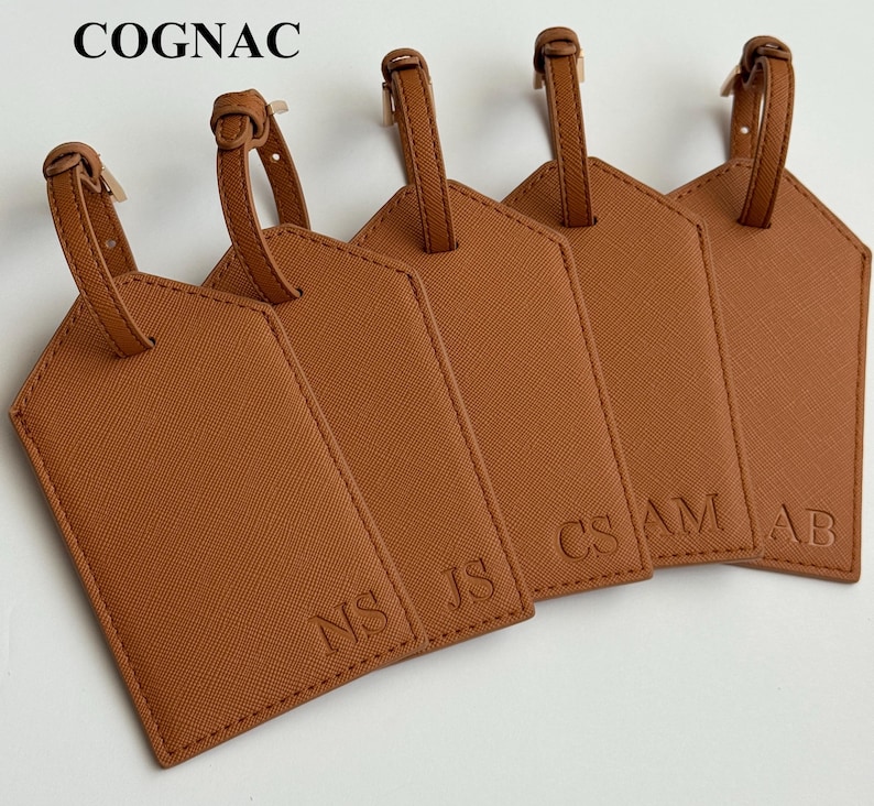 May include: Five cognac-colored luggage tags with matching straps. Each tag has a different set of initials embossed on the front: NS, JS, CS, AM, and AB. The word "COGNAC" is printed at the top.